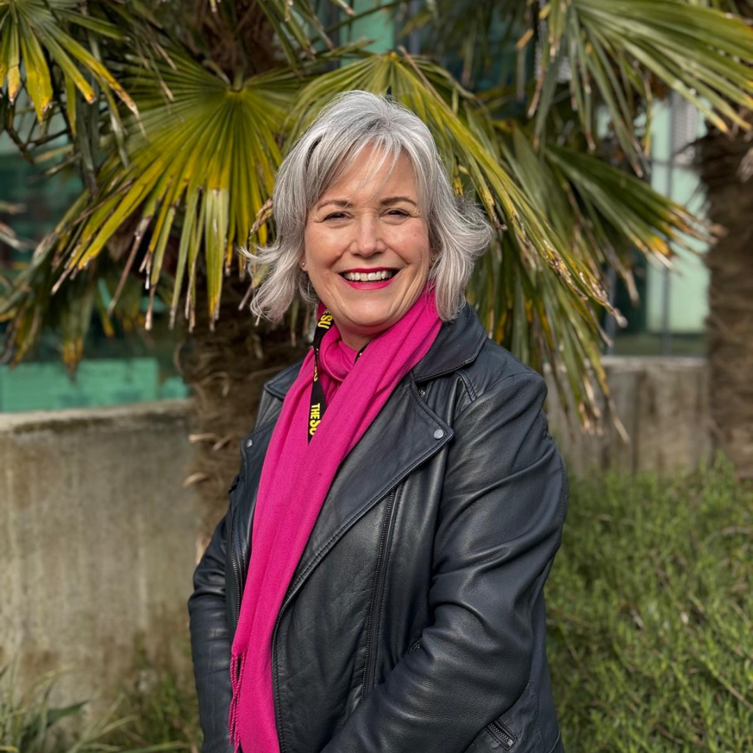 Angie smiles at the camera while stood in front of palm trees on Penryn Campus
