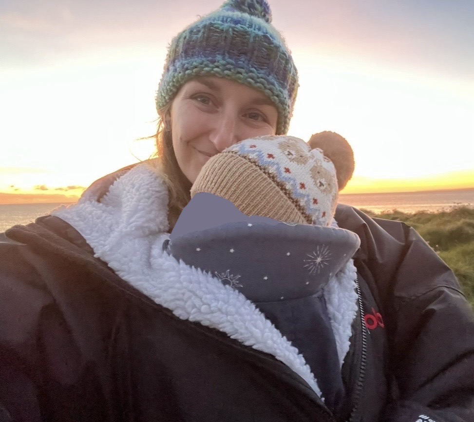 Georgi wears a beanie hat and smiles at the camera over the head of her small baby who is wrapped up in a baby carrier. She has blonde wavy hair.