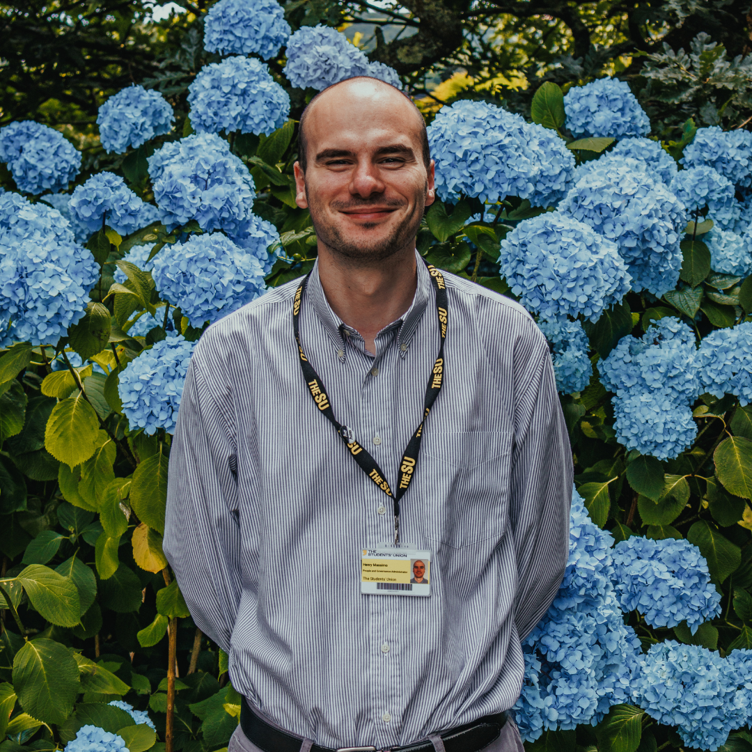 Henry is wearing a striped shirt and smiling at the camera. He is stood in front of blue flowers