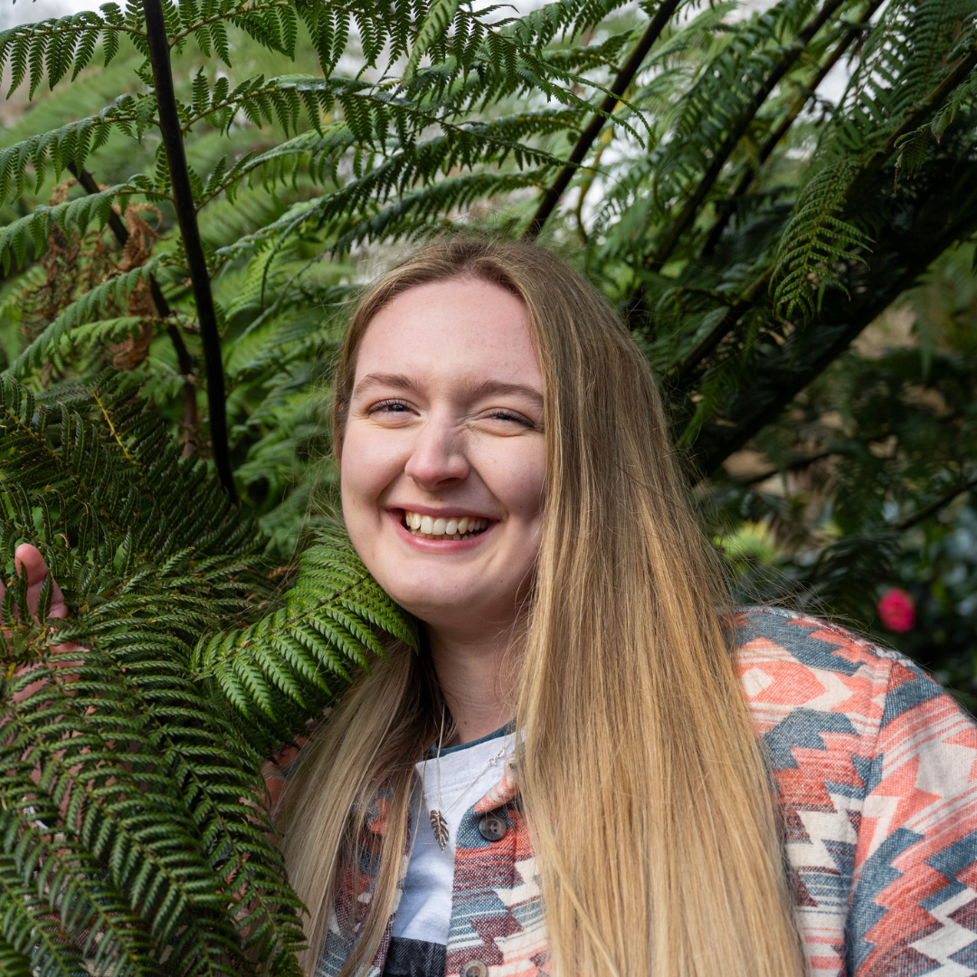 Photo shows Izzy smiling with a large plant by her