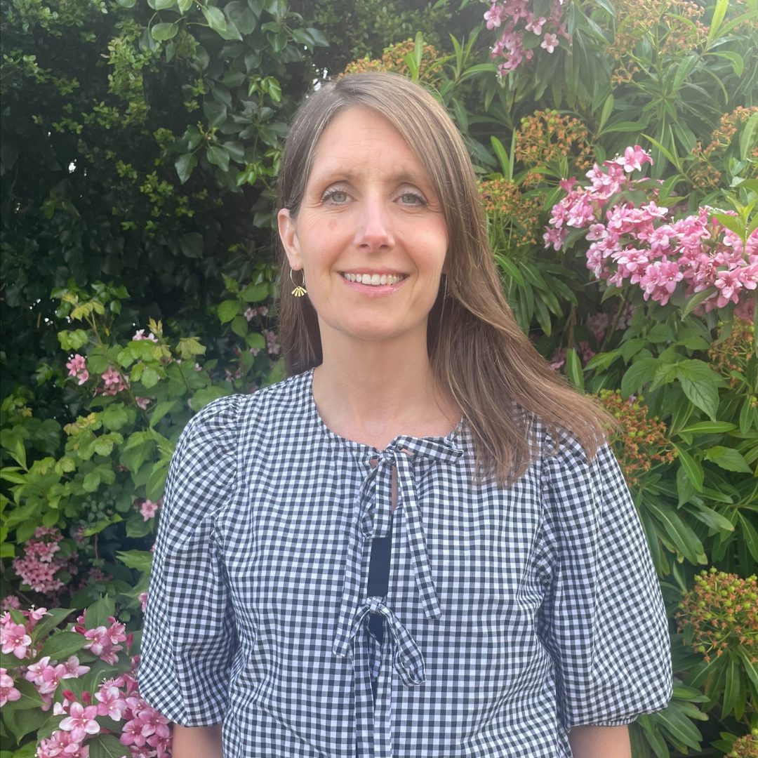 Jo stands in front a green plant with flowers. She has long brown hair and wears and checked top with bows on the front.