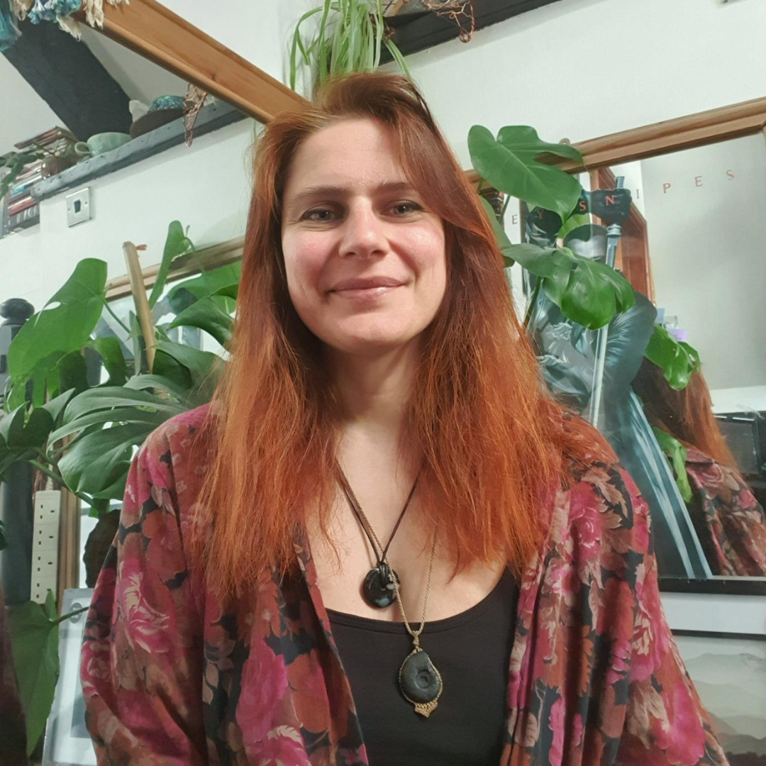 Katie, a white woman smiles, facing the camera directly. She wears a black top and red cardigan with a chunky long necklace.