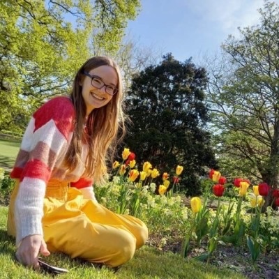 Penny, a white woman wearing glasses, sits in a garden next to red and yellow tulips. She smiles at the camera as the sun shines on her brown hair. She wears a red jumper and yellow trousers that perfectly match the tulips.