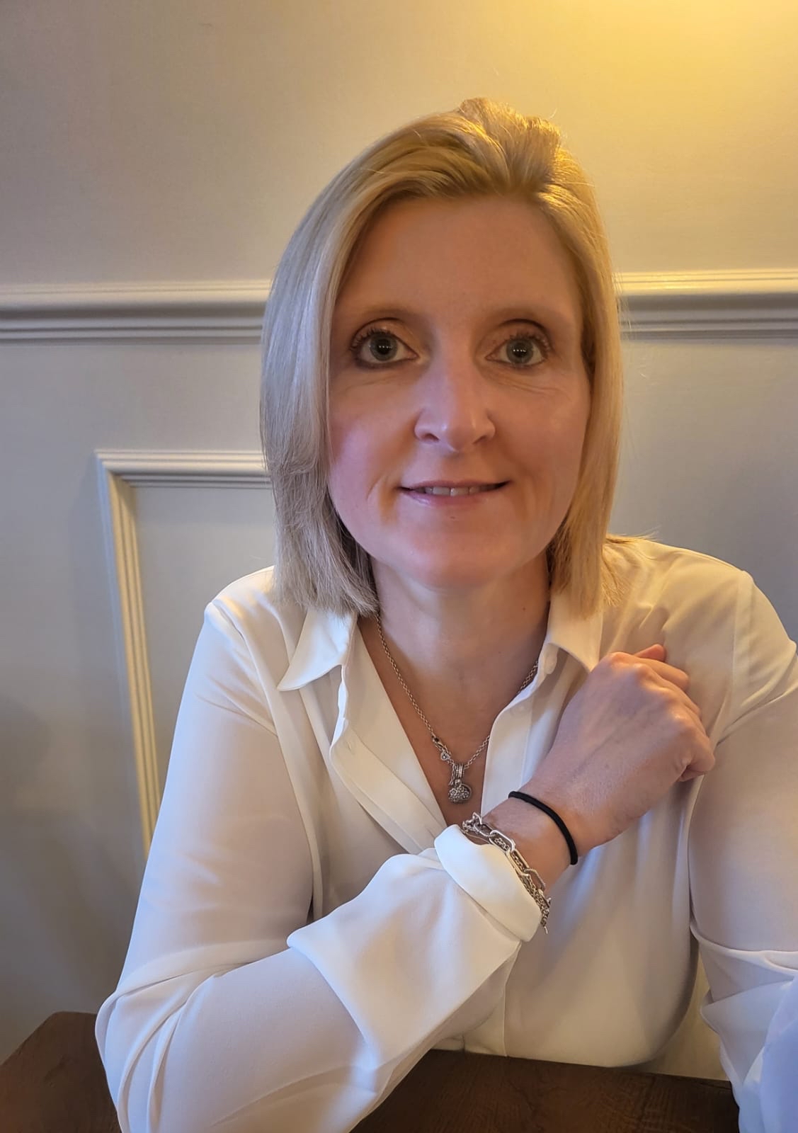 Rhian is a white woman with blode hair to her chin. She smiles at the camera and wears a white shirt and silver jewellery.