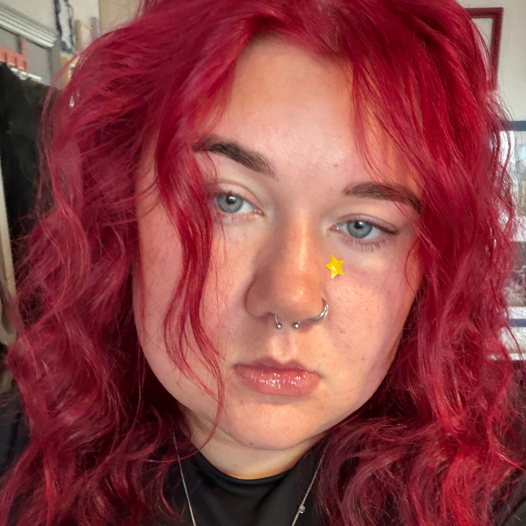 Bethan has bright red hair, blue eyes and a nose ring and septum piercing. she is wearing a yellow star pimple patch an staring at the camera