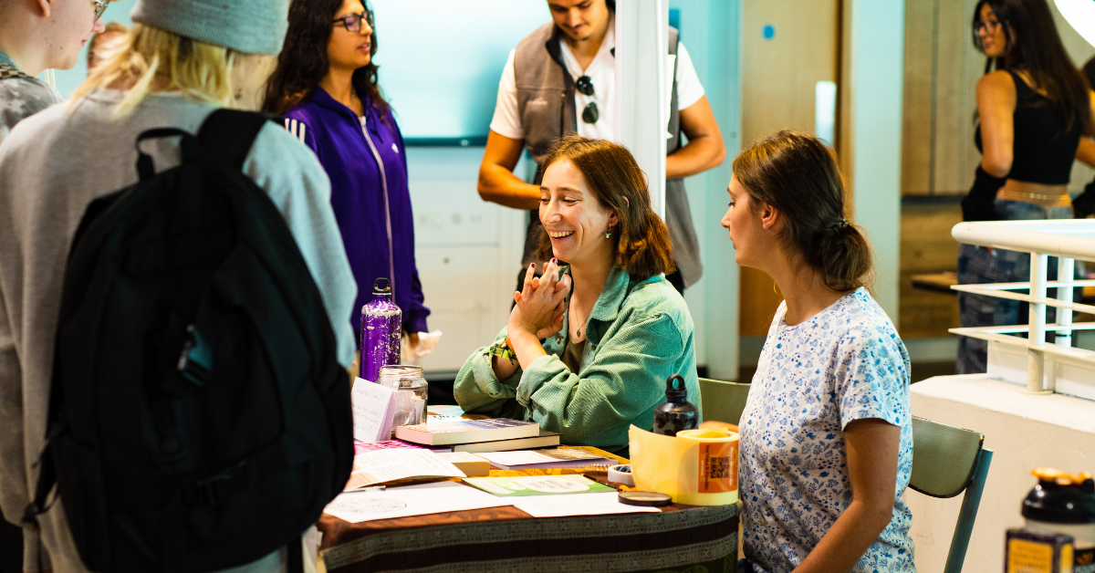 Two committee members sit at a table and talk to students interested in joining their society.