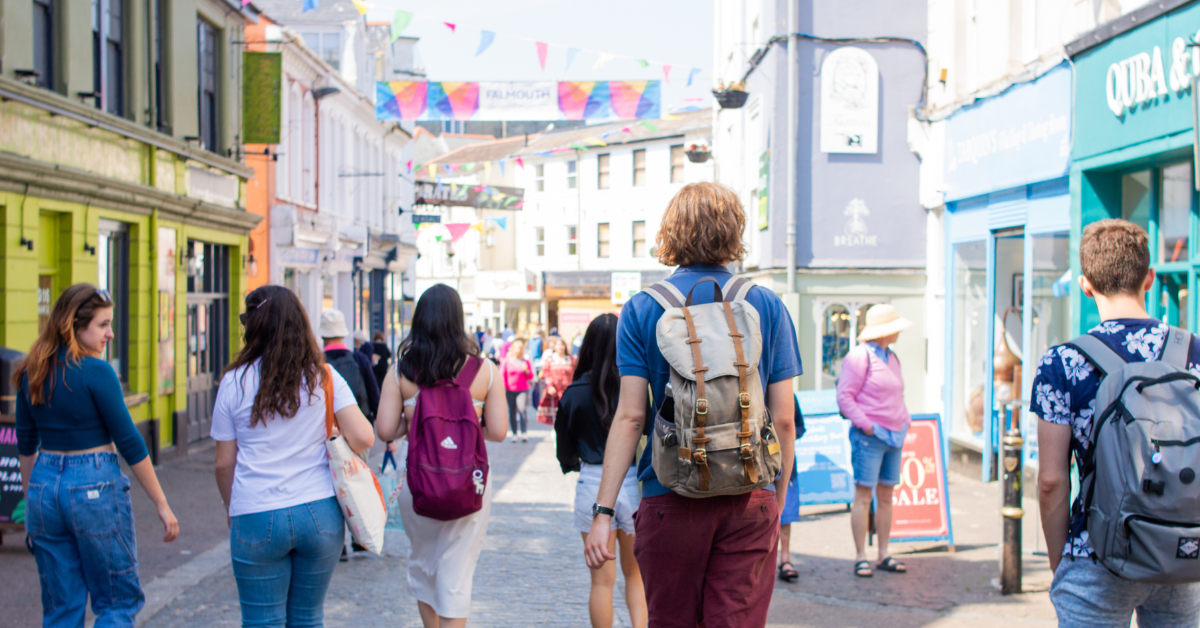Students walking down Falmouth Town High Street on a sunny day. Some are wearing backpacks, and others are carrying bags as they explore the area decorated with vibrant bunting overhead.